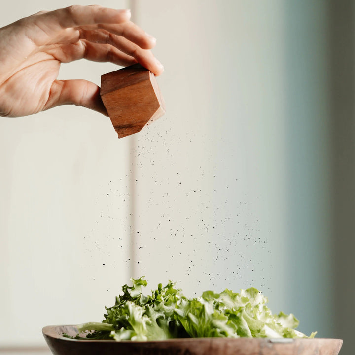 Hand holding a NORDICO acacia wooden salt or pepper shaker, sprinkling seasoning over a fresh green salad in a wooden bowl.