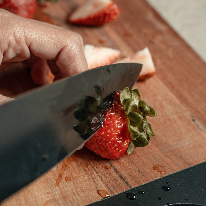 Close-up of a Nordico black-handled knife slicing a strawberry on an acacia wood cutting board.