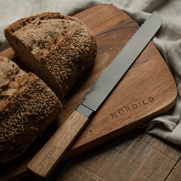 Nordico serrated stainless steel knife with acacia handle slicing bread on a natural wood cutting board.