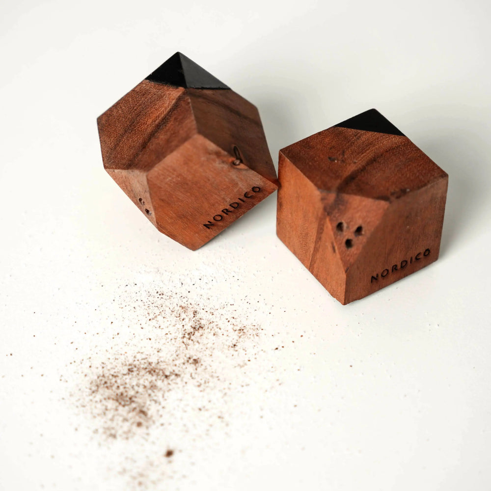Two NORDICO salt and pepper shakers made of premium acacia wood with black hand-painted accents, displayed on a white surface with scattered pepper.