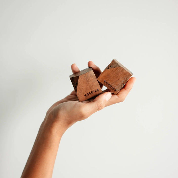 Person holding a pair of NORDICO acacia wooden salt and pepper shakers in hand, showcasing their geometric shape and engraved branding.