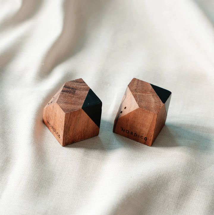 Close-up of two NORDICO acacia wood salt and pepper shakers with black geometric accents, placed on beige fabric.