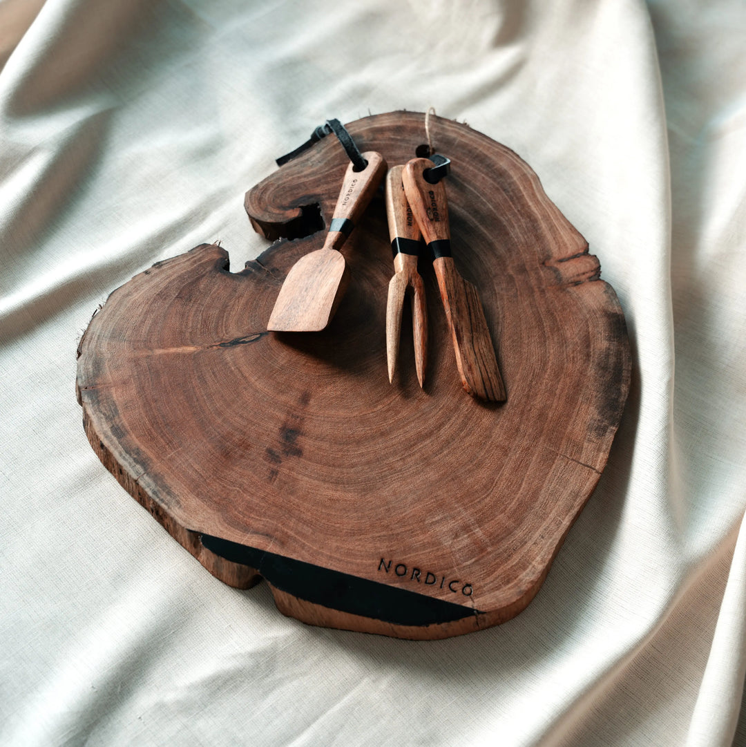 Nordico Cheese Board with Utensils (Stylized Shot) – Rustic acacia wood cheese board with cheese knives placed artfully, showcasing its irregular and unique natural shape.