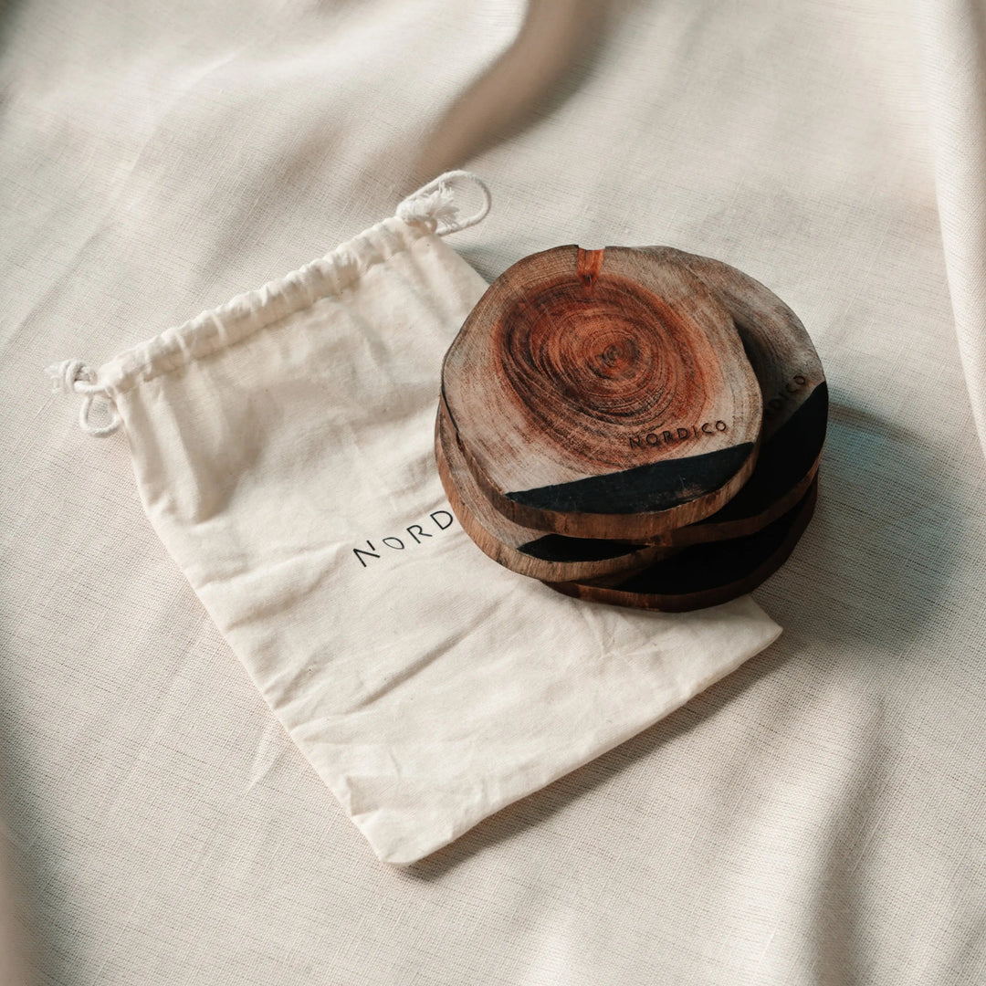 NORDICO Acacia wooden coasters set stacked on a beige fabric background next to a cotton drawstring storage pouch with the Nordico logo.