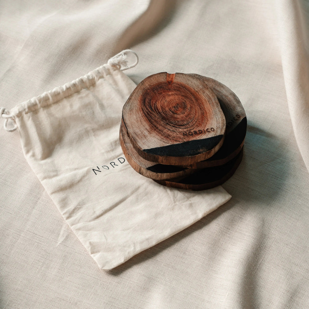 Close-up of NORDICO Acacia wooden coasters stacked neatly beside a branded cotton drawstring pouch, displaying wood grain patterns.