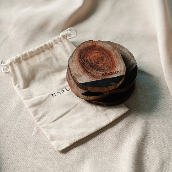 Close-up of NORDICO Acacia wooden coasters stacked neatly beside a branded cotton drawstring pouch, displaying wood grain patterns.