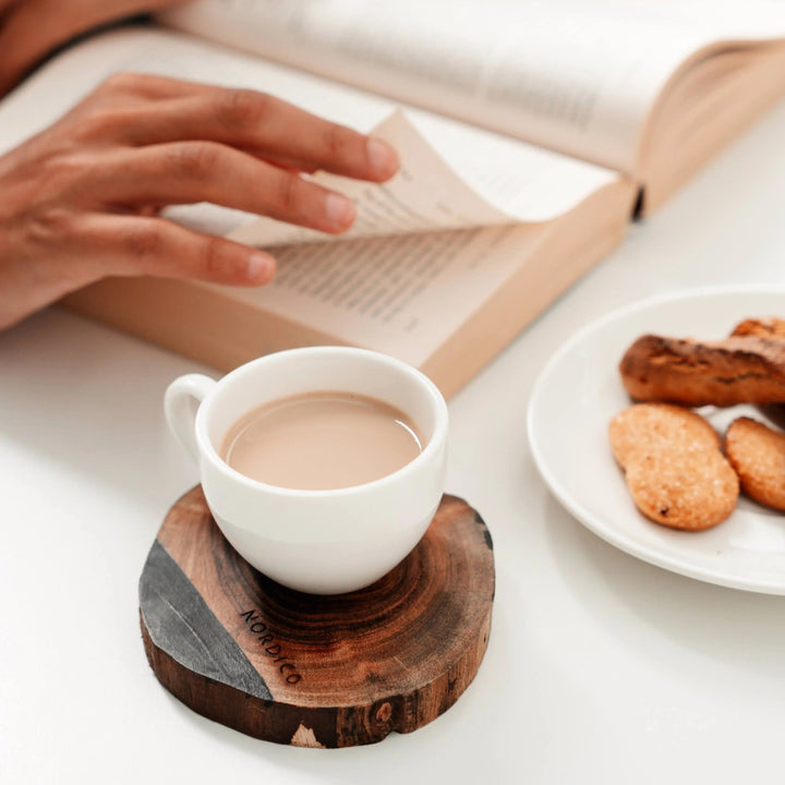 NORDICO wooden coaster holding a white coffee cup with tea or coffee, placed next to a book and a plate of cookies, creating a cozy setup.