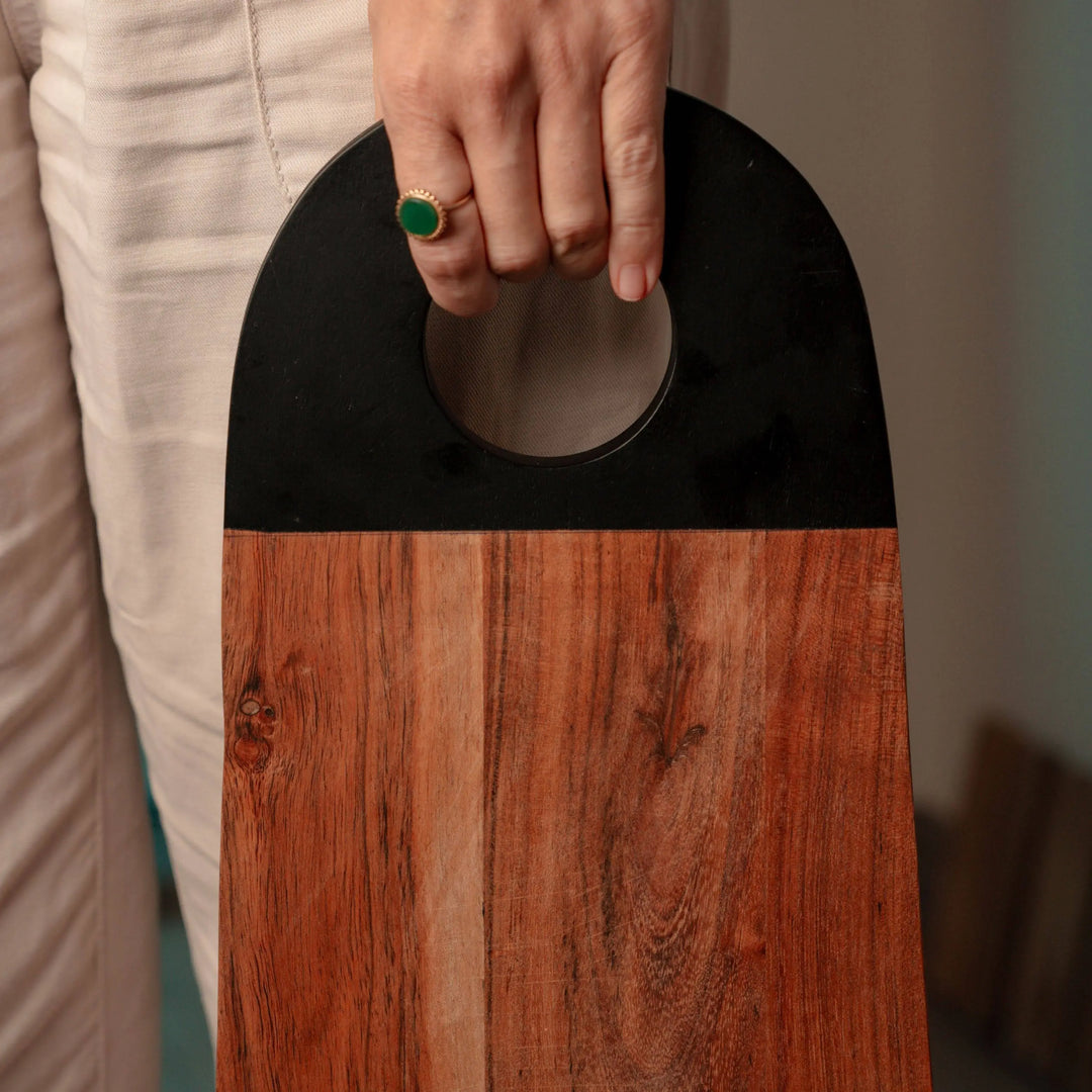Person holding a Nordico acacia wood cutting board by its black-painted handle, displaying its ergonomic design.