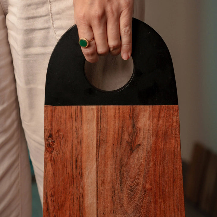 Person holding a Nordico acacia wood cutting board by its black-painted handle, displaying its ergonomic design.