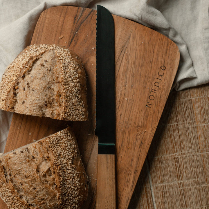 Nordico acacia cutting board with a black bread knife and sliced artisan bread, perfect for kitchen tasks and serving.