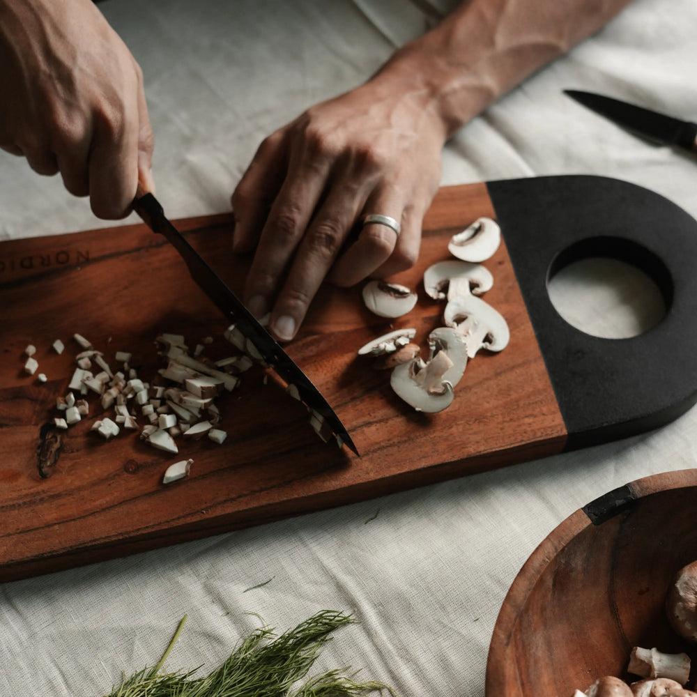 Person chopping mushrooms on a Nordico acacia cutting board with black-painted handle in a kitchen setting.
