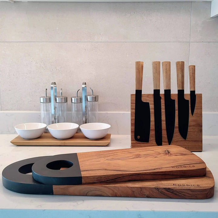 Nordico medium-sized acacia cutting board with a black handle, styled with kitchen accessories on a white countertop.